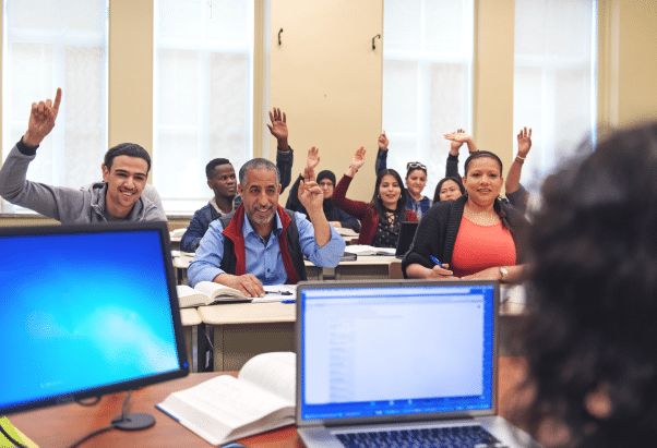 A diverse group of adult learners seated in a classroom, smiling and raising their hands to participate. The foreground shows an open laptop and a textbook on a desk, suggesting an interactive and engaging learning environment.