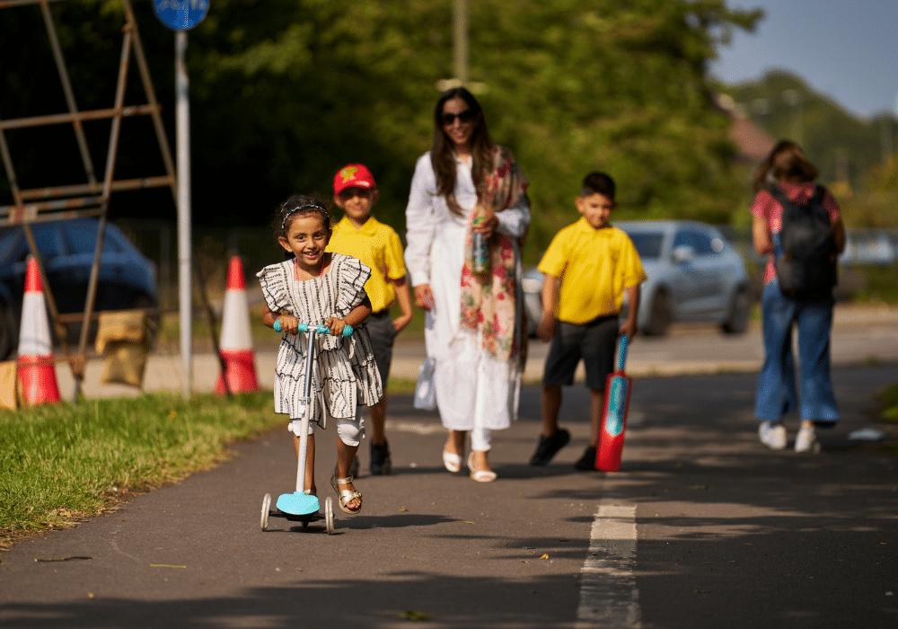 A young girl rides her scooter on a path. Her family are walking behind her.