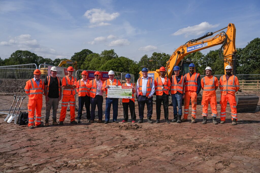 Twelve people wearing high-visibility orange safety clothing and hard hats stand together on a construction site. Mayor Claire stands in the middle posing with a project sign in front of a large excavator. Several ceremonial spades are visible to the left, with temporary fencing and trees in the background.