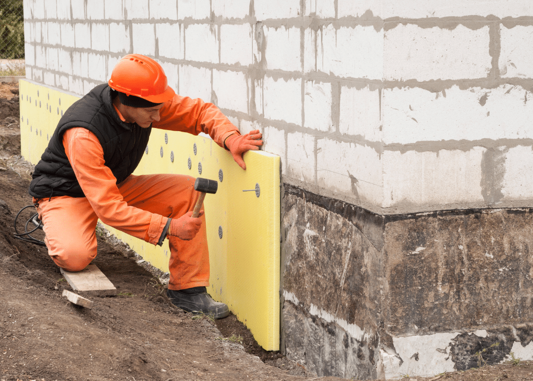 A person in high vis and hard hat is hammering insulation panels on to the outside of a building.